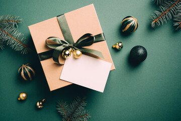 Elegant Christmas gift box with ribbon bow, blank paper note, balls decoration and fir branches on green background. Vintage, retro style Xmas present, New year surprise concept.