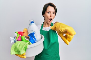 Young brunette woman with short hair wearing apron holding cleaning products pointing with finger surprised ahead, open mouth amazed expression, something on the front