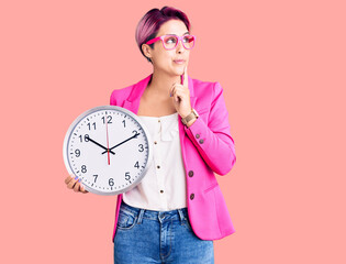 Young beautiful woman with pink hair wearing business jacket and holding clock serious face...