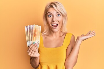 Obraz premium Young blonde girl holding norwegian krone banknotes celebrating achievement with happy smile and winner expression with raised hand