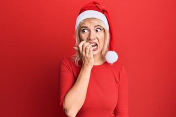 Young blonde girl wearing christmas hat looking stressed and nervous with hands on mouth biting...