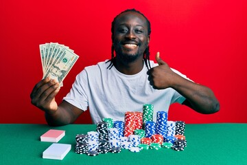 Handsome young black man playing poker holding 20 dollars banknotes smiling happy and positive, thumb up doing excellent and approval sign