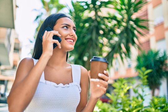Young latin girl talking on the smartphone drinking take away coffee at street of city.