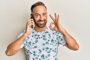 Fototapeta premium Handsome middle age man having conversation talking on the smartphone doing ok sign with fingers, smiling friendly gesturing excellent symbol