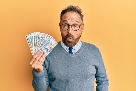 Handsome middle age man holding dollars scared and amazed with open mouth for surprise, disbelief face