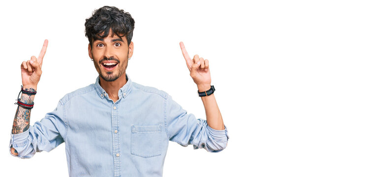 Young hispanic man wearing casual clothes smiling amazed and surprised and pointing up with fingers and raised arms.