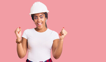 Young african american woman with braids wearing hardhat and builder clothes screaming proud,...