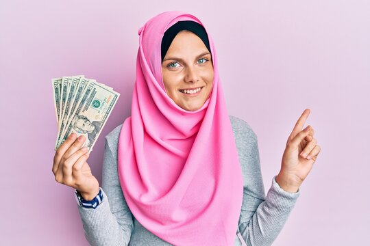 Young caucasian woman wearing islamic hijab holding 20 dollars banknotes smiling happy pointing with hand and finger to the side