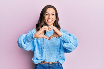 Young brunette woman wearing casual winter sweater over pink background smiling in love doing heart symbol shape with hands. romantic concept.