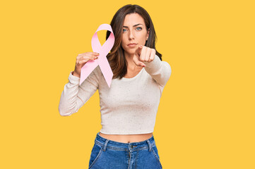 Young brunette woman holding pink cancer ribbon pointing with finger to the camera and to you, confident gesture looking serious