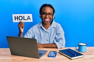 Young african woman wearing operator headset holding hola word looking positive and happy standing and smiling with a confident smile showing teeth © Krakenimages.com