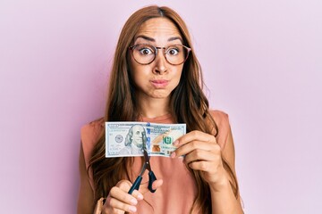 Young brunette woman cutting dollars with scissors for currency devaluation puffing cheeks with funny face. mouth inflated with air, catching air.