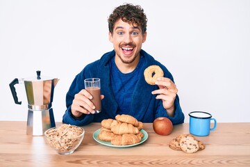 Young caucasian man with curly hair eating breakfast holding chocolate beverage and donut smiling and laughing hard out loud because funny crazy joke.