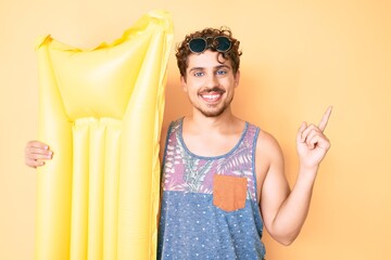 Obraz premium Young caucasian man with curly hair wearing summer clothes and holding matress float smiling happy pointing with hand and finger to the side