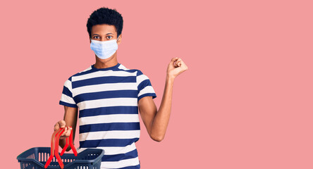 Young african american man wearing shopping basket and medical mask screaming proud, celebrating victory and success very excited with raised arms