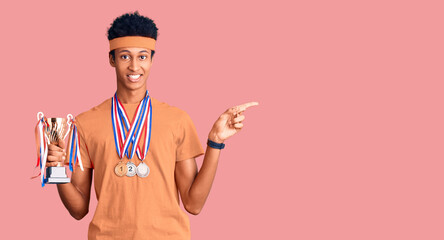 Obraz na płótnie Canvas Young african american man holding champion trophy wearing medals smiling happy pointing with hand and finger to the side
