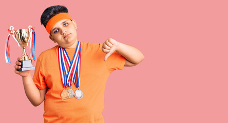 Little boy kid holding champion trophy and wearing medals with angry face, negative sign showing dislike with thumbs down, rejection concept