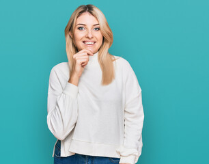 Young caucasian woman wearing casual winter sweater looking confident at the camera smiling with crossed arms and hand raised on chin. thinking positive.
