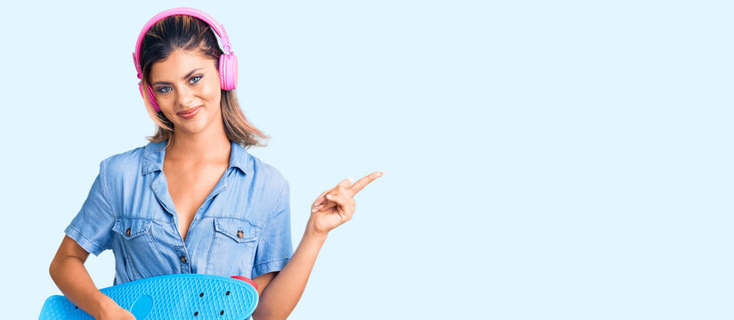 Young beautiful woman wearing headphones and holding skate smiling happy pointing with hand and finger to the side