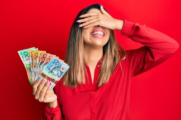 Beautiful brunette woman holding australian dollars smiling and laughing with hand on face covering eyes for surprise. blind concept.