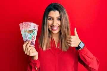 Beautiful brunette woman holding 100 new zealand dollars banknote smiling happy and positive, thumb up doing excellent and approval sign