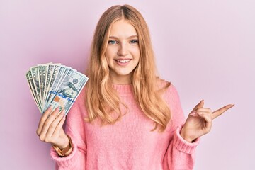 Beautiful young caucasian girl holding dollars smiling happy pointing with hand and finger to the side