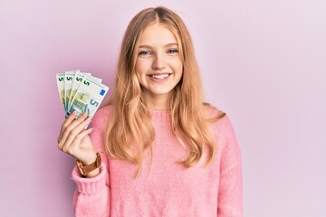 Beautiful young caucasian girl holding bunch of 5 euro banknotes looking positive and happy standing and smiling with a confident smile showing teeth