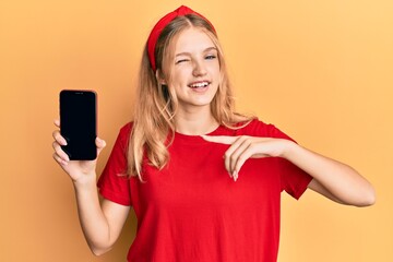 Beautiful young caucasian girl holding smartphone showing screen winking looking at the camera with...