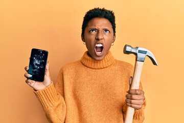 African american woman with short hair holding broken smartphone holding hammer winking looking at the camera with sexy expression, cheerful and happy face.