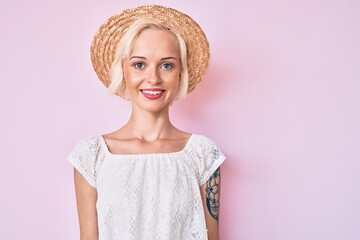 Young blonde woman with tattoo wearing summer hat with a happy and cool smile on face. lucky person.