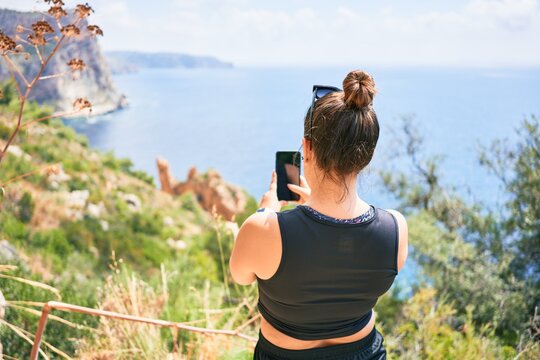 Young hispanic woman on back view wearing sportswear. Making photo using smartphone standing over beautiful landscape