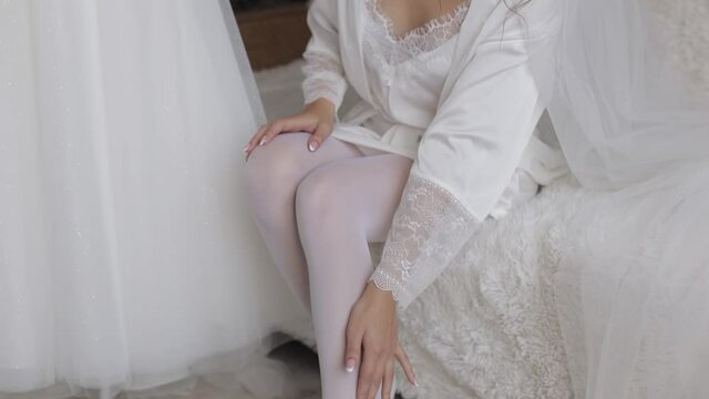 Slim legs of young bride wearing stylish white lacy rest gown and high heeled shoes sitting on bed in bedroom. Woman put on wearing wedding shoes. Bridal footwear, morning dressing before ceremony