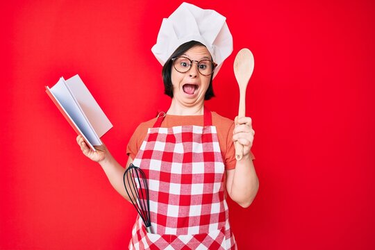 Brunette woman with down syndrome wearing professional baker apron reading cooking recipe book celebrating crazy and amazed for success with open eyes screaming excited.
