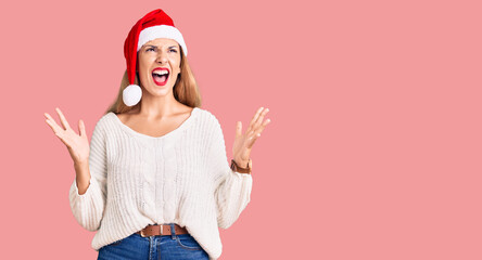 Fototapeta premium Beautiful young woman wearing christmas hat crazy and mad shouting and yelling with aggressive expression and arms raised. frustration concept.