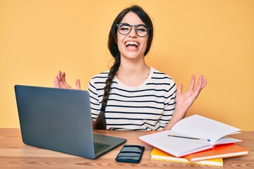 Brunette teenager girl working at the office with laptop celebrating mad and crazy for success with arms raised and closed eyes screaming excited. winner concept