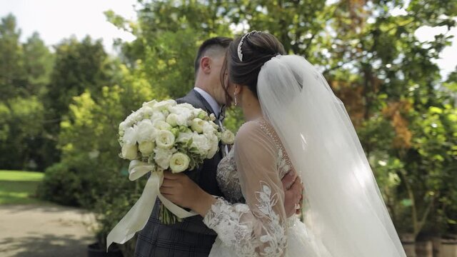 Newlyweds. Lovely young caucasian bride and groom making a kiss in park. Wedding family couple. Man and woman in love. Summer. Bride in gorgeous luxury wedding dress. Bridegroom in jacket. Slow motion