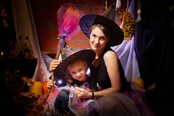 Obraz na płótnie Canvas Beautiful brunette mother and cute little daughter looking as witches in special dresses and hats in room decorated for Halloween. Witchcraft and wizardry in carnival. Halloween style photo shoot.
