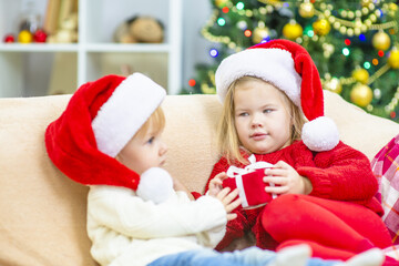 Little girls are sitting in Santa hats at home on the couch against the background of a Christmas tree and giving each other a gift. Cozy holidays concept