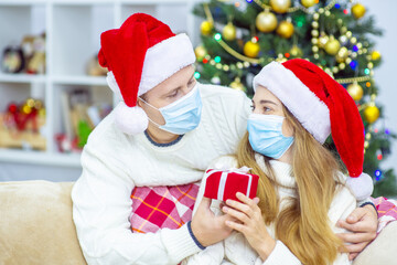 Young married couple with medical masks wearing santa hats with a gift in their hands wish each other a Merry Christmas (New Year)