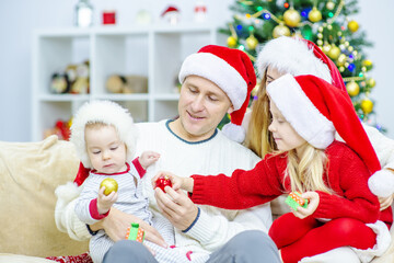 A big friendly family of four is sitting together on a sofa against the background of a Christmas tree and looking at the very youngest child. Cozy holidays at home concept