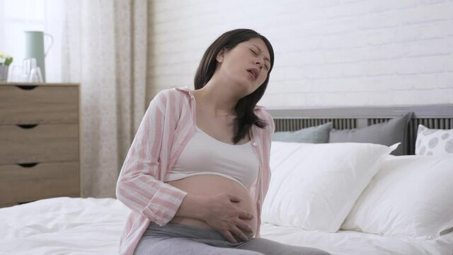 asian expectant mother suffering labor contraction is moaning and holding her tummy while her face is contorted in a grimace of pain in the bedroom.