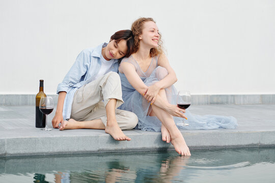 Happy smiling yuong Asian woman leaning on shoulder of girlfriend and closing eyes with pleasure when couple resting by swimming pool