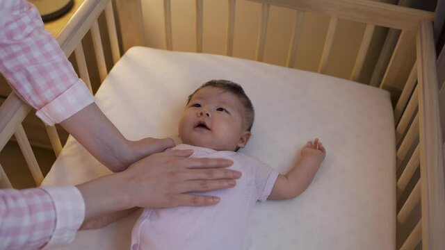 portrait adorable asian female infant in baby cot looking up at her mother and babbling while being patted in the bedroom at home.