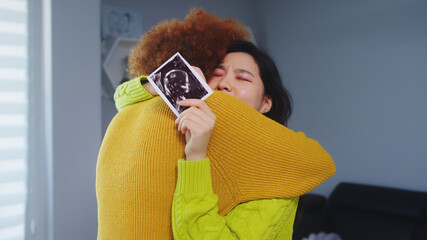 Multiracial lesbian couple expecting baby. Embracing with ultrasound baby picture. Maternity...