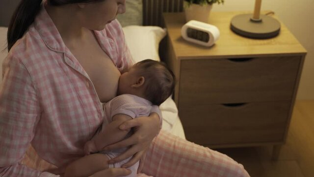 high angle shot Taiwanese first time mom cuddling and gazing lovingly while breastfeeding her baby daughter to sleep at bedside.