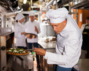 Skilled male chef in white uniform working in professional kitchen of restaurant, baking ordered pizza
