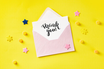 Hand written Thank You card in a pink envelope, overhead shot on a yellow background with glitter stars and candies
