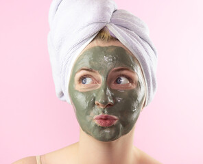 A young woman with a towel on her head and a clay mask on her face on a pink background. Face care, Spa treatments at home.