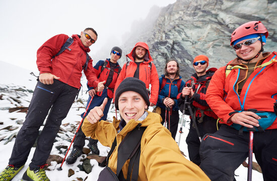 Young man taking picture with friends hikers during travel in winter mountains. Group of male travelers looking at camera and smiling, showing thumbs up. Concept of travelling, hiking and photography.