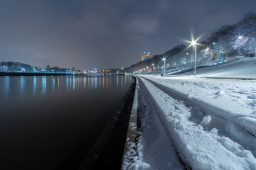 View of the Moskva River embankment at night in winter. The stone steps of the embankment, sidewalks and trees are covered with fresh snow. The backlight is on on the shore. Colorful city landscape.